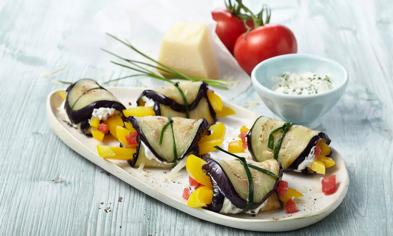 Gerollte Auberginenscheiben mit Frischkäsefüllung, garniert mit gelber Paprika und Schnittlauch, auf ovaler heller Platte angerichtet