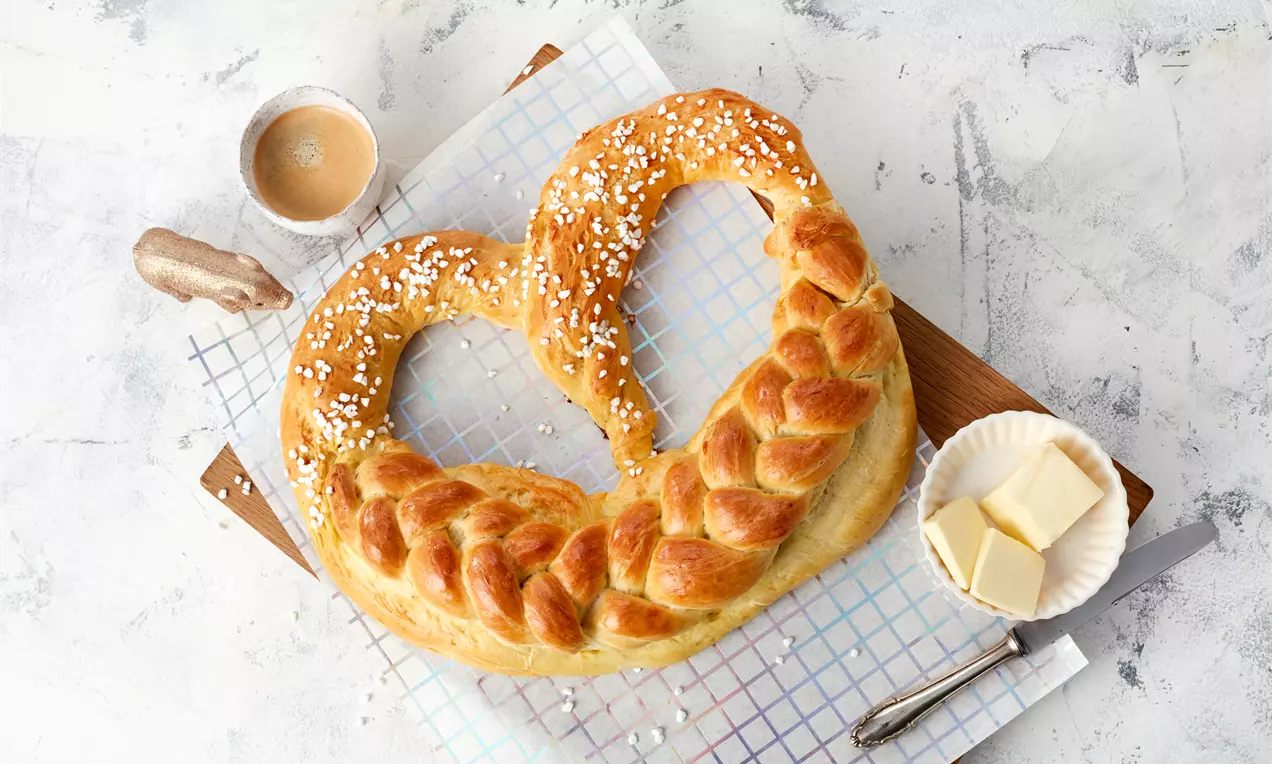 Goldbraune, glänzende Neujahrsbrezel mit grobem Hagelzucker, herzförmig und flechtartig, auf einem Holzbrett mit Butterstücken und Kaffee