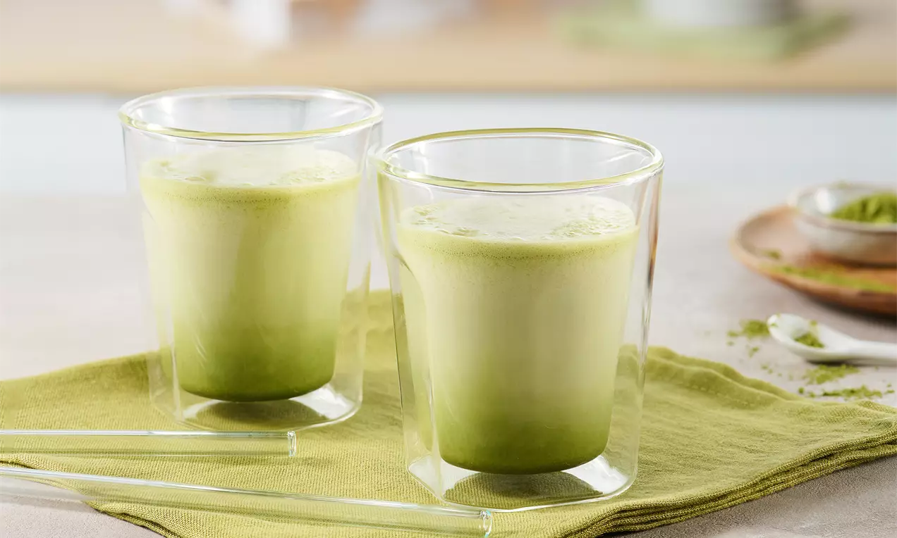 Zwei Gläser Matcha Latte mit sanftem Farbverlauf von kräftigem Grün zu cremigem Hellgrün auf grünem Tuch, daneben Glasstrohhalme und Pulver