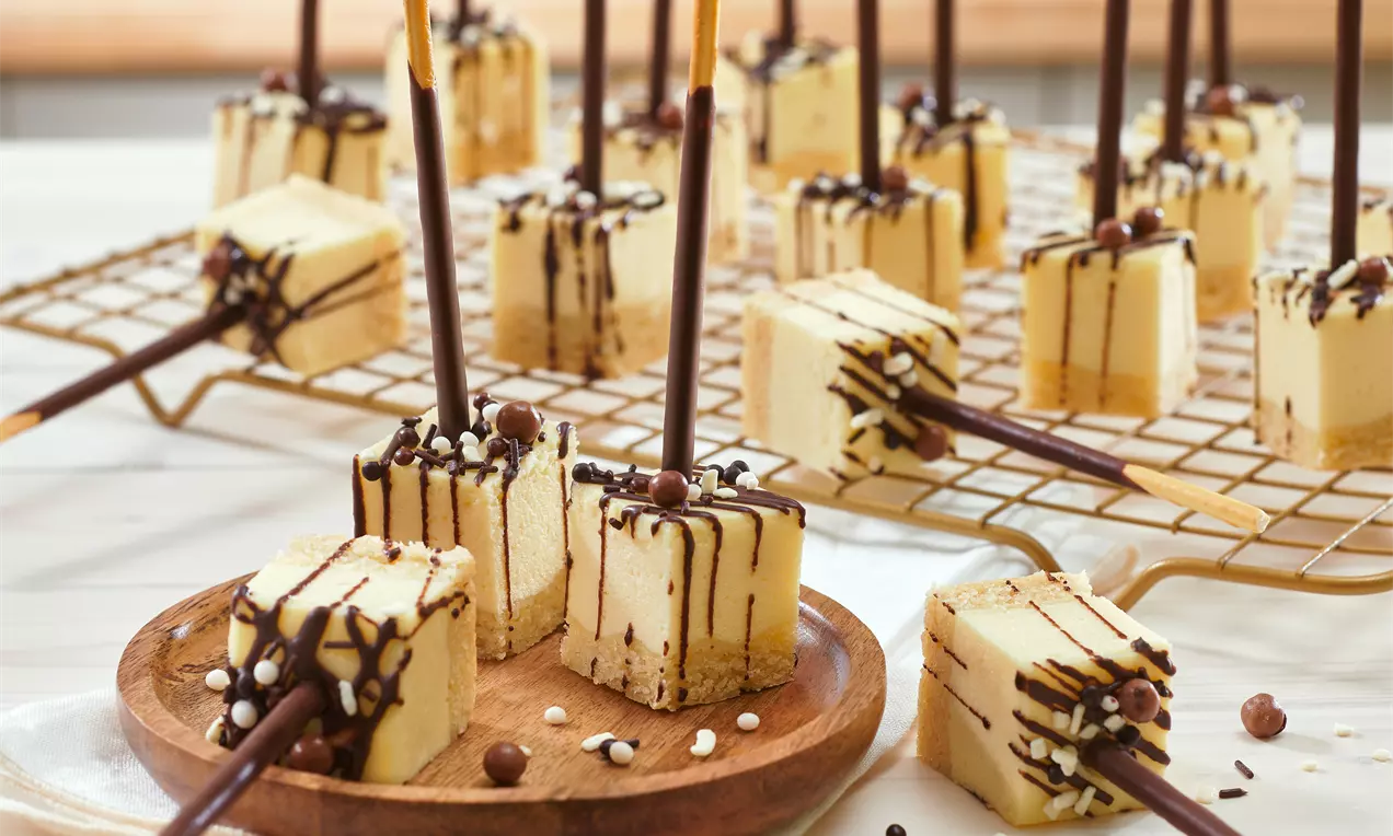 Quadratische Käsekuchen-Happen mit hellem Keksboden, gelber Creme und dunkler Schokoladendekoration, arrangiert mit Schokoladensticks