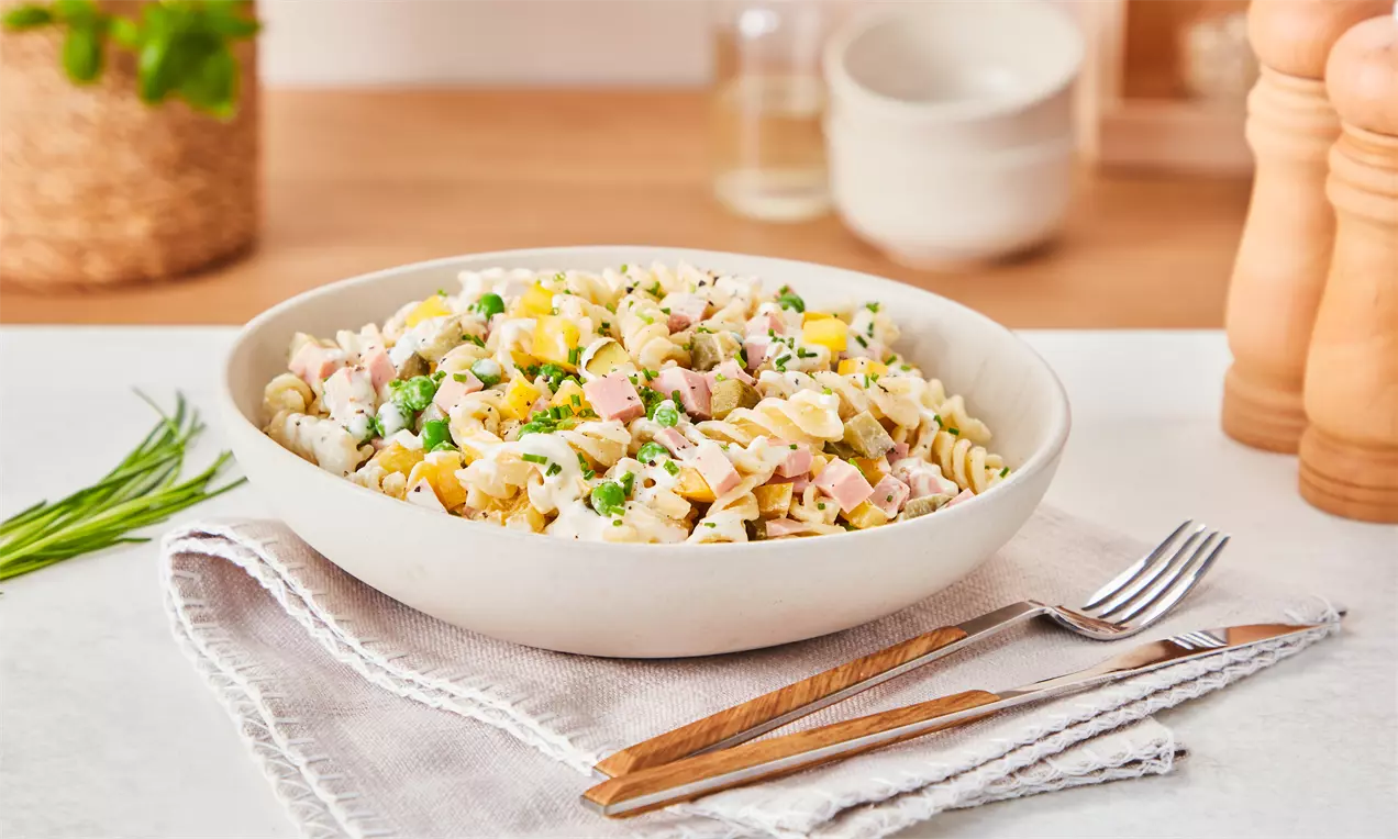 Bunt gemischter Nudelsalat mit gelbem Mais, pinkem Schinken, grünen Erbsen und cremigem Dressing in einer weißen Schüssel auf Stoffservietten