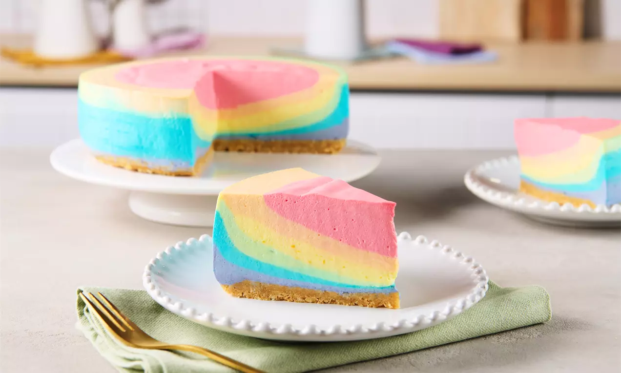 Regenbogenfarbiger Cheesecake mit geschichteten Pastelltönen auf Keksboden, serviert auf weißem Teller mit goldener Gabel auf hellgräner Serviette