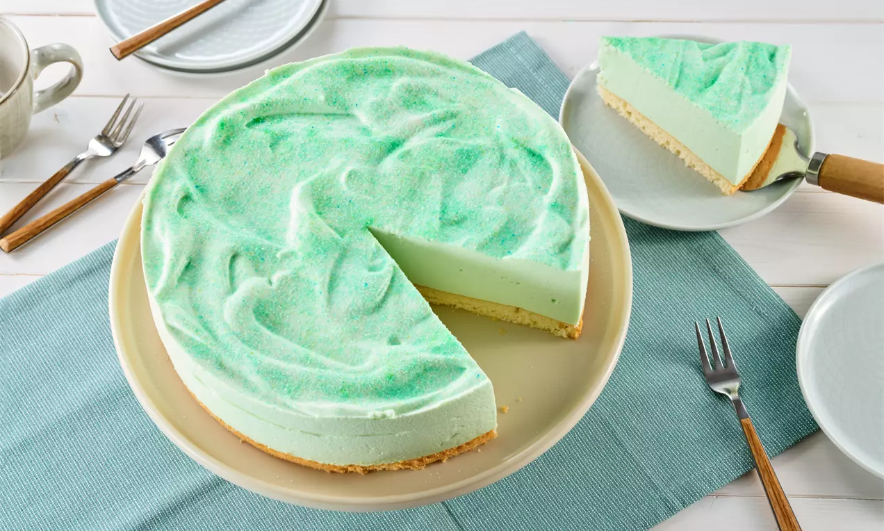 Waldmeistertorte mit leicht grün geschwungener Creme auf hellem Keksboden, ein angeschnittenes Stück auf einem Teller daneben angerichtet