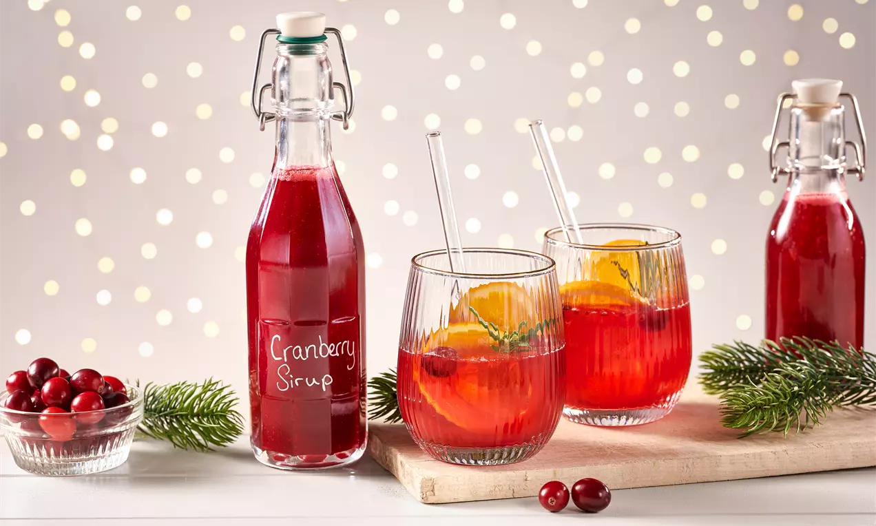 Glasflasche mit dunkelrotem Cranberrysirup neben gläsern mit orange- und rosmarindekoration auf hellem holzbrett mit tannenzweigen