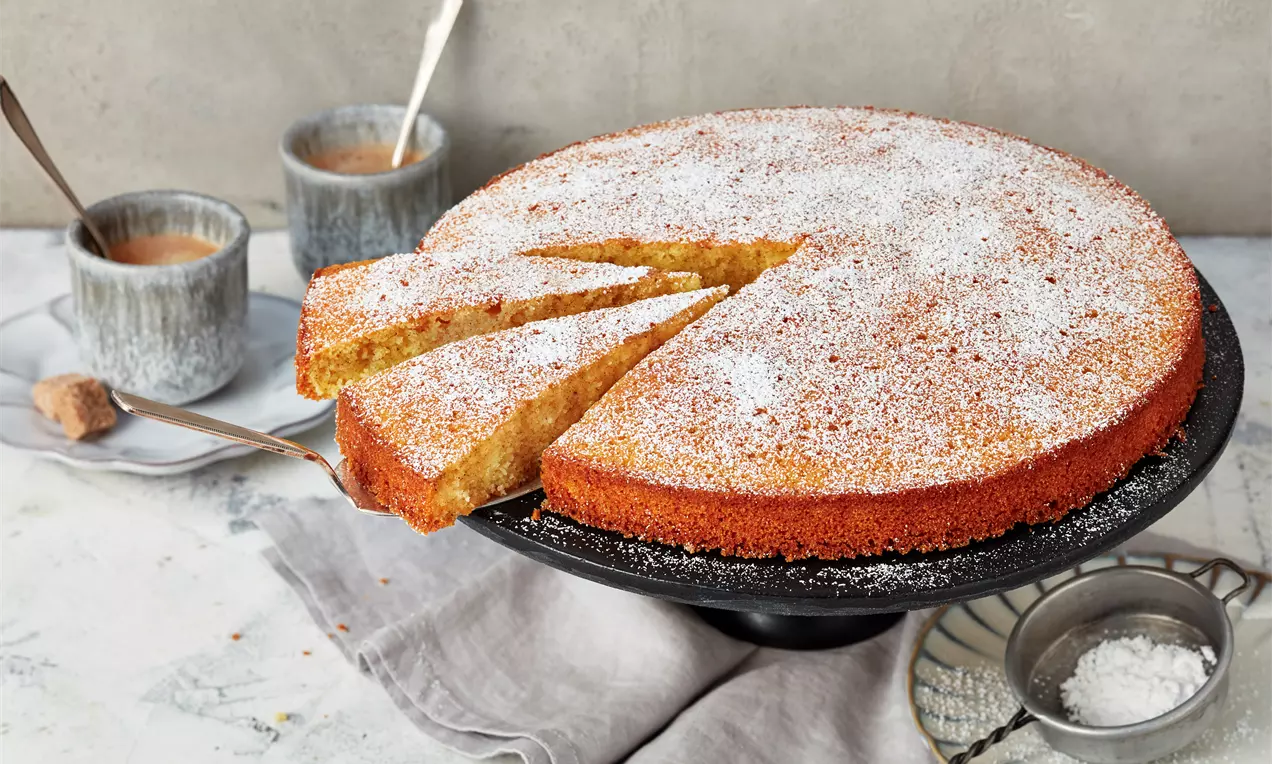 Goldbrauner, leichter Grießkuchen mit leicht knuspriger Oberfläche und feinem Puderzucker, serviert auf schwarzer Kuchenplatte mit grauen Tassen