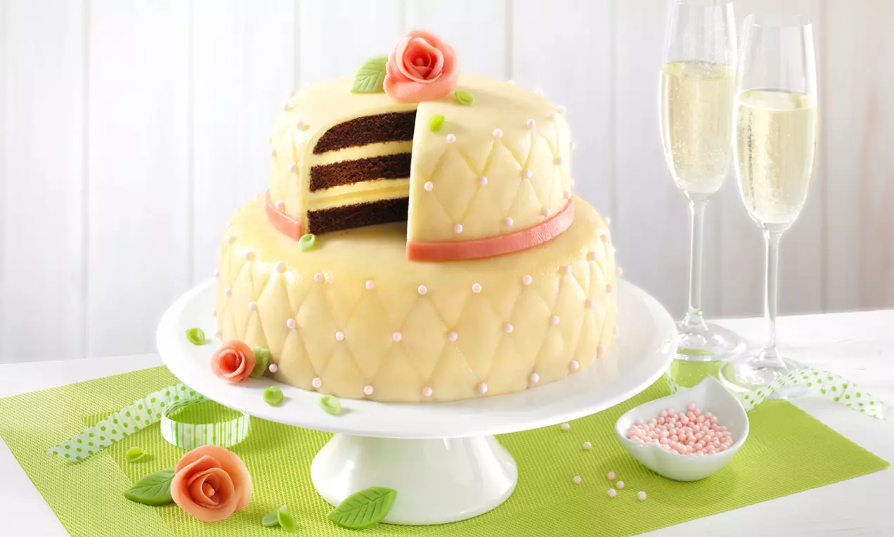 Zweistöckige Marzipantorte mit cremefarbener Oberfläche, rosa Blüten, zartem Muster und einem angeschnittenen Stück auf weißer Tortenplatte