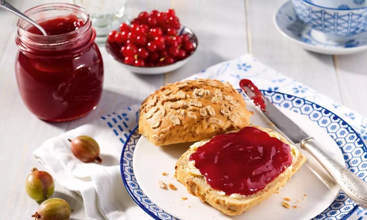 Samtig glänzende Stachelbeer-Johannisbeer-Konfitüre in tiefroter Farbe auf einem knusprigen Brötchen, mit Früchten und Glas dekoriert