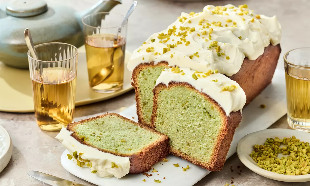 Aufgeschnittener Pistazienkuchen mit hellgrüner Krume, weißer Creme und gehackten Pistazien auf einer Platte, serviert mit Tee und Pistazienstückchen