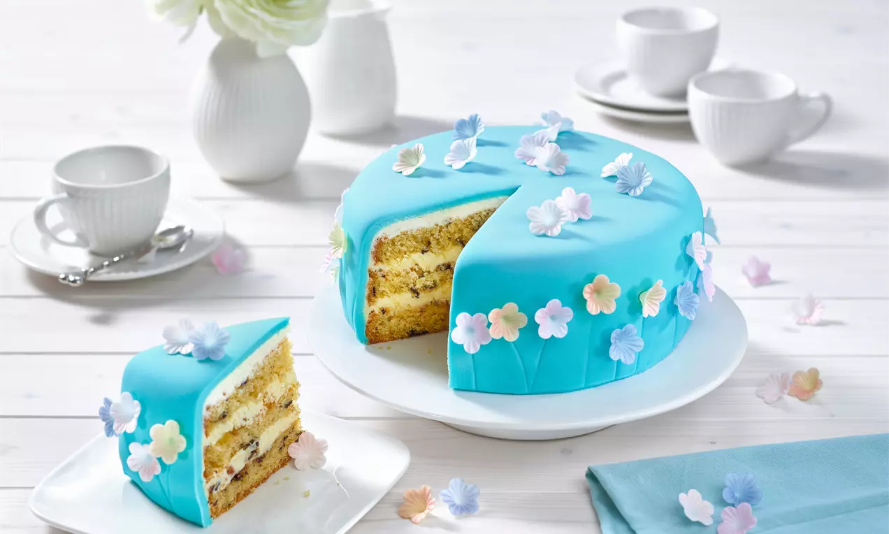 Türkise Torte mit geschichteter Füllung, dekoriert mit pastellfarbenen Blüten, auf weißer Tortenplatte mit passendem Anschnittstück