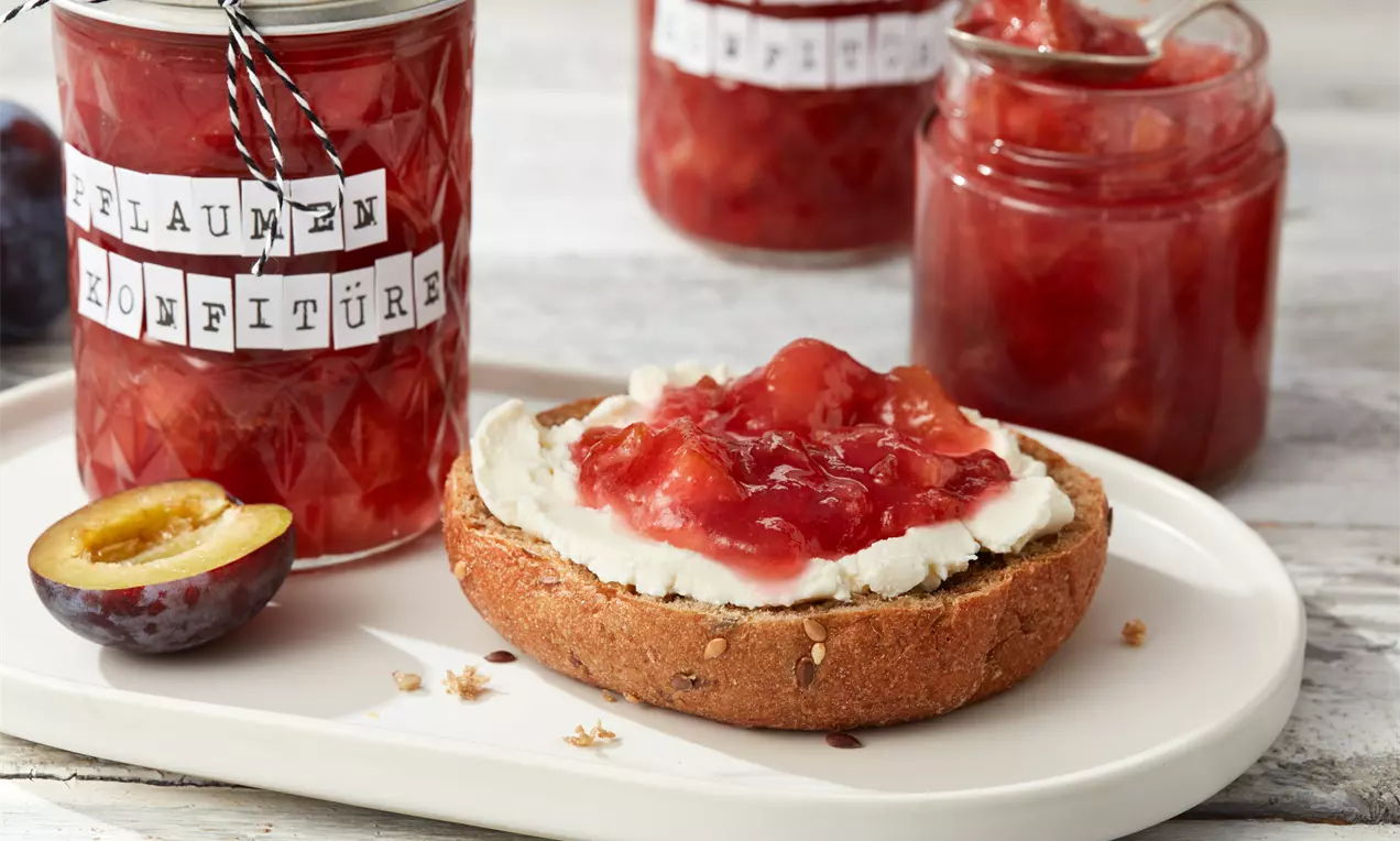 Pflaumenkonfitüre in einem geöffneten und einem geschlossenen Glas neben einer aufgeschnittenen dunkelroten Pflaume und einem mit Frischkäse bestrichenen Brötchen