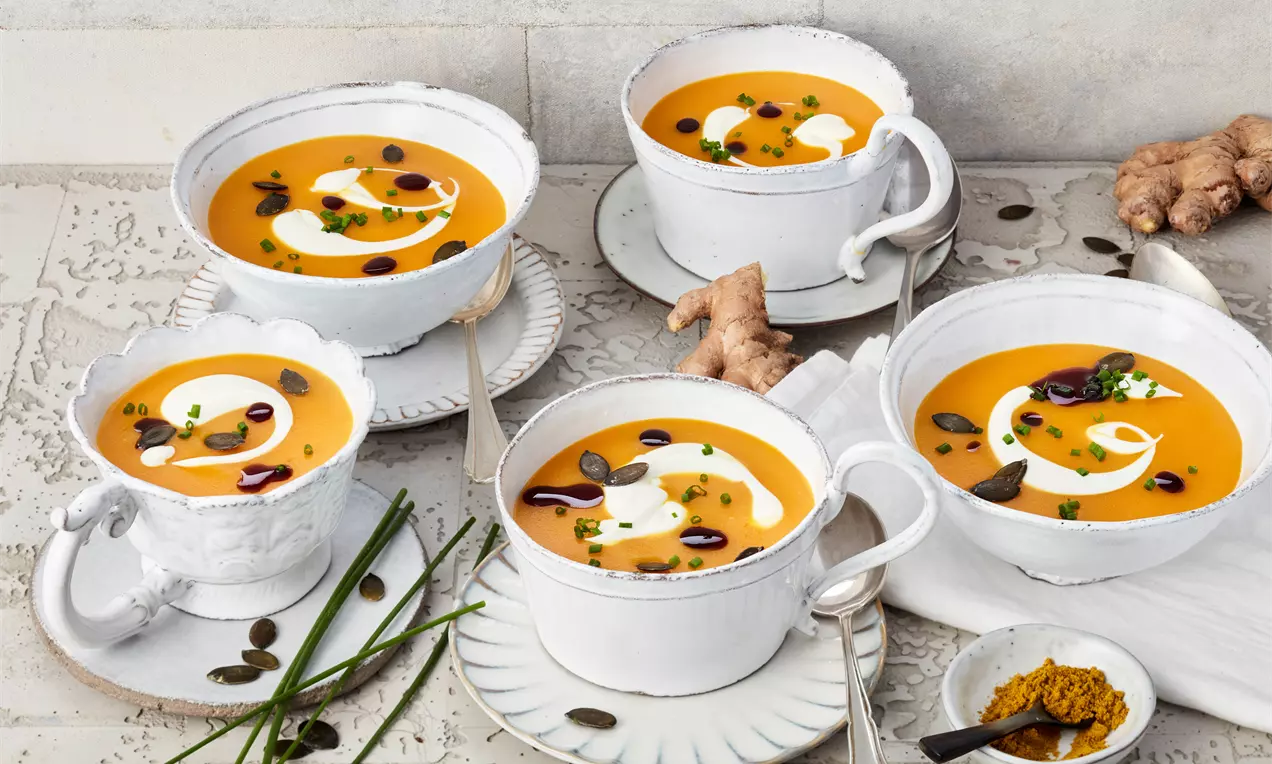 Cremig orangefarbene Butternut-Kürbis-Suppe mit weißem Sahne-Swirl, Kürbiskernen und Kräutern in weißen Tassen auf heller Steinplatte