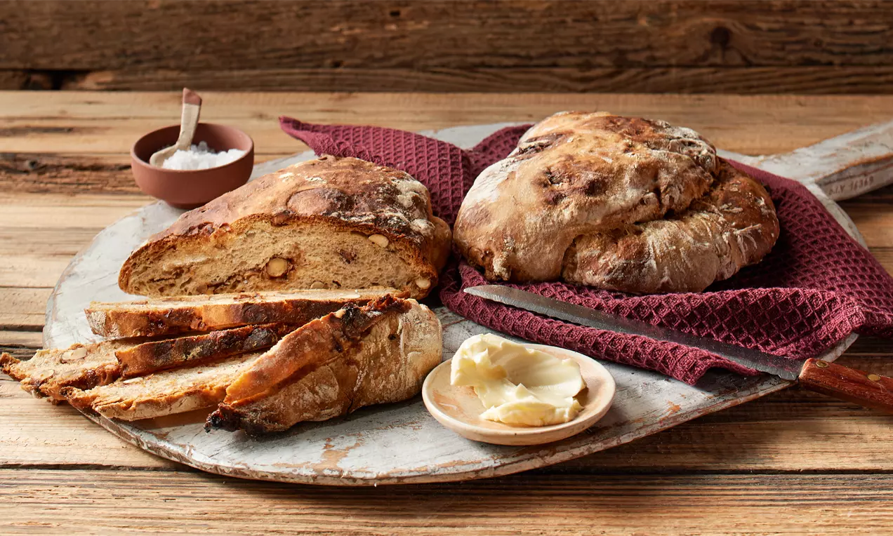 Rustikales Haselnussbrot mit brauner Kruste, Scheiben und Laib auf Holzbrett mit rotem Tuch, Butter und Salz in kleinen Gefäßen