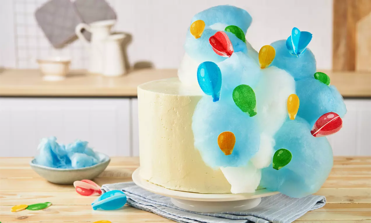 Helle Torte mit glatter Buttercreme, dekoriert mit blauer Zuckerwatte und bunten Luftballon-Zuckerfiguren auf weißem Teller mit Stoffunterlage