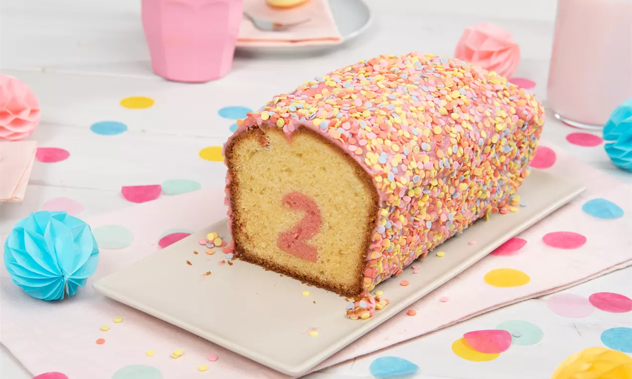 Rosa Überraschungskuchen mit bunter Zuckerstreuselglasur und rosa Zahl im Inneren auf weißer Platte auf dekorativem Tisch mit Pastellfarben