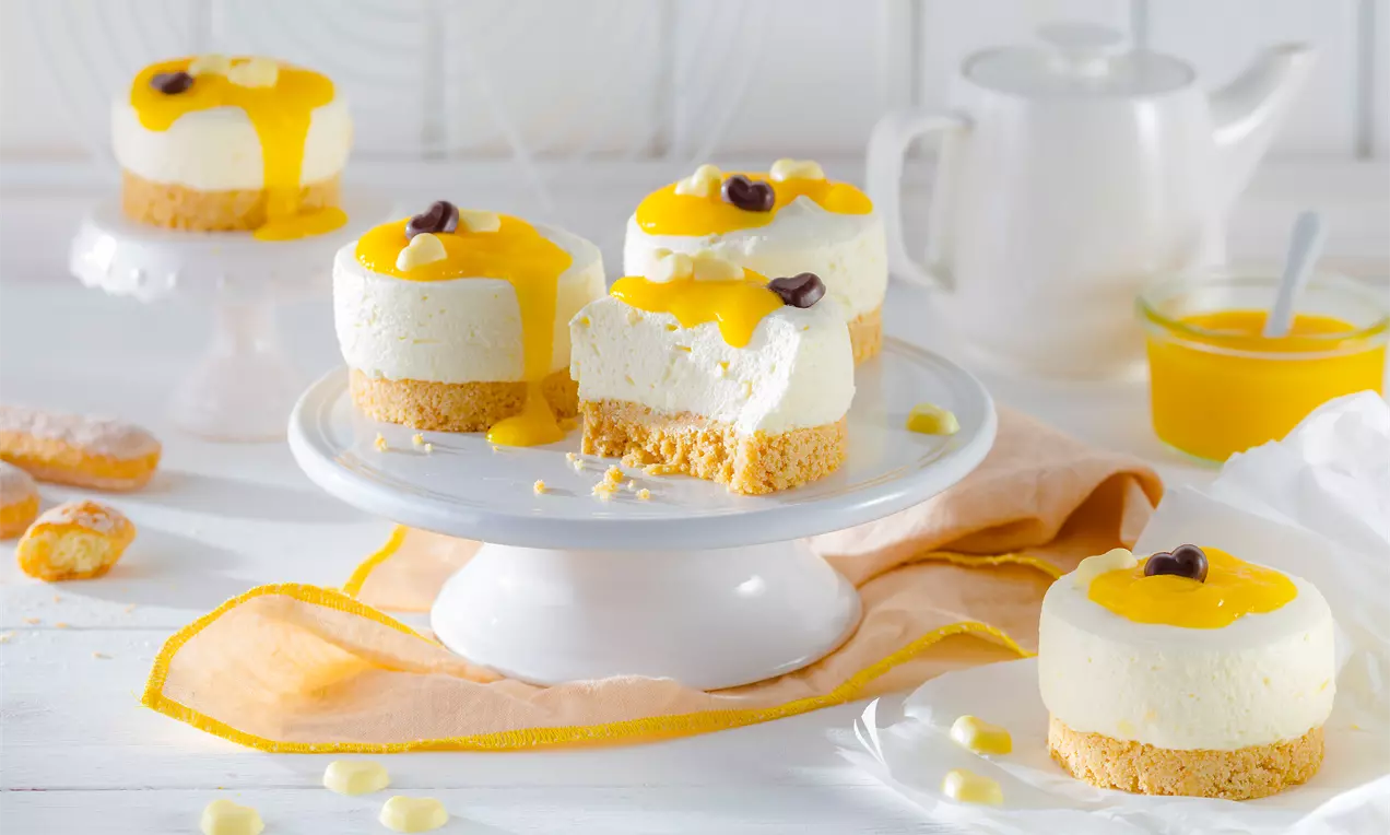 Kleine weiße Mousse-Törtchen auf Keksboden mit gelber Mangosoße, garniert mit Schoko-Deko, arrangiert auf weißem Teller mit Mangosoße als Beilage