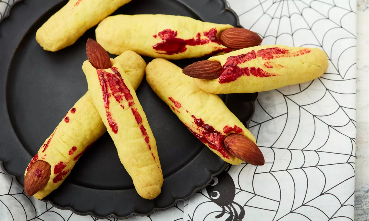 Hexenfinger aus goldbraunem Gebäck mit Mandelnägeln und roter Marmelade als 'Blut', auf rustikalem Blech und Backpapier angerichtet