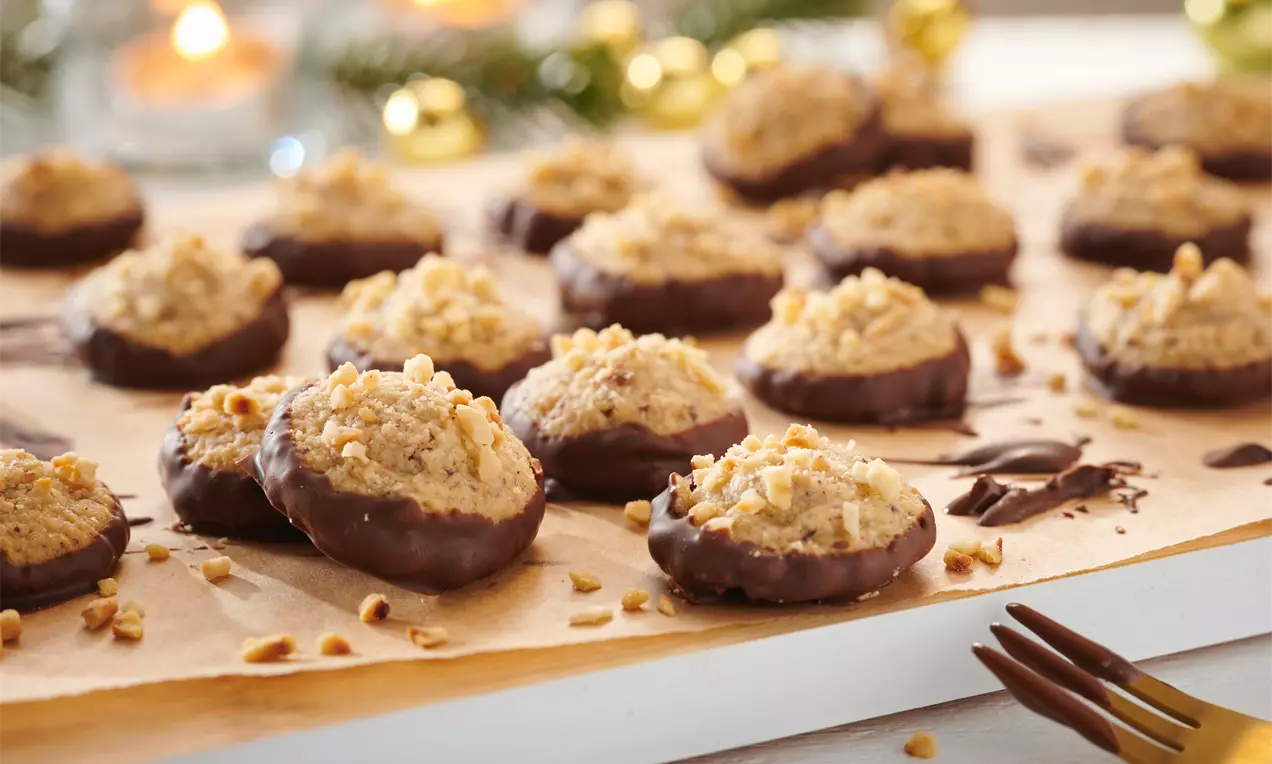 Marzipan-Makronen mit goldbraunem Haselnusskern und dunkler Schokoglasur auf Backpapier, umgeben von dekorativen Weihnachtslichtern