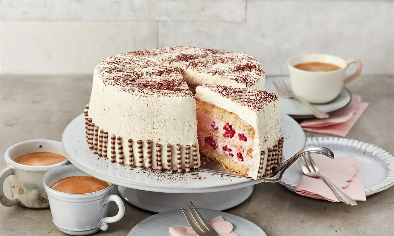 Helle Himbeer-Sahnetorte mit lockerer Füllung, Schokoladendekor und Kakao bestäubt, angeschnitten auf einer weißen Tortenplatte mit Kaffee serviert