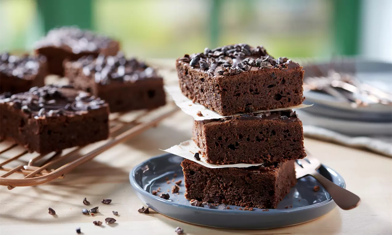 Dunkelbraune, rechteckige Brownie-Stücke mit lockerer Textur, gestapelt auf grauem Teller, bestreut mit Kakao-Nibs und Backpapier dazwischen
