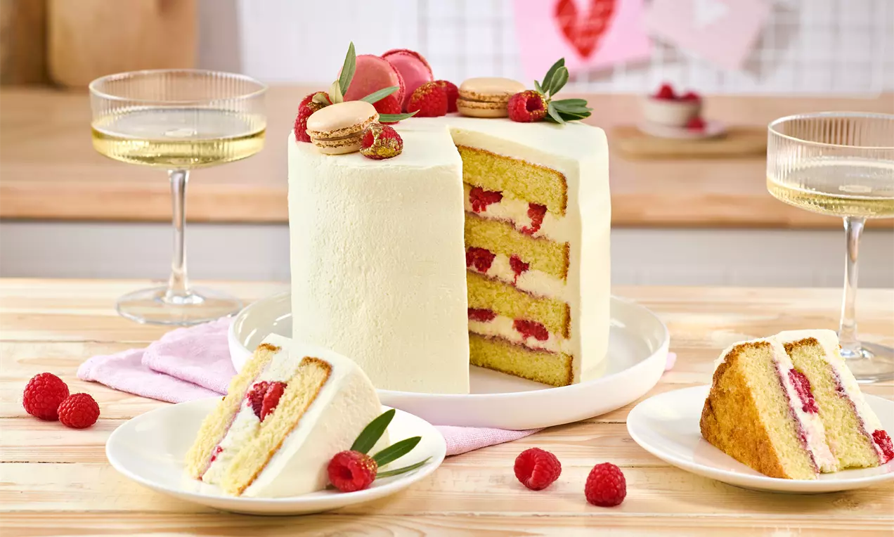Hohe moderne Hochzeitstorte mit weißer Creme, gefüllt mit Himbeeren und Sahneschichten. Oben dekoriert mit Himbeeren, Macarons und Grün. Zwei angeschnittene Stücke liegen davor.