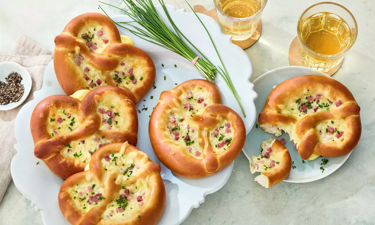 Flammkuchenbrezeln mit goldbraunem Rand, cremigem Belag, Speckwürfeln und Schnittlauch auf weißer Platte, daneben Gläser mit Apfelschorle