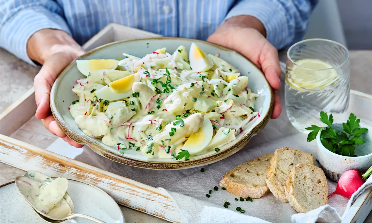 Ein Teller mit Spargelsalat aus weißen Spargelstücken, Radischenscheiben, gekochten Eier-Vierteln und Schnittlauch, präsentiert auf einem Holztablett