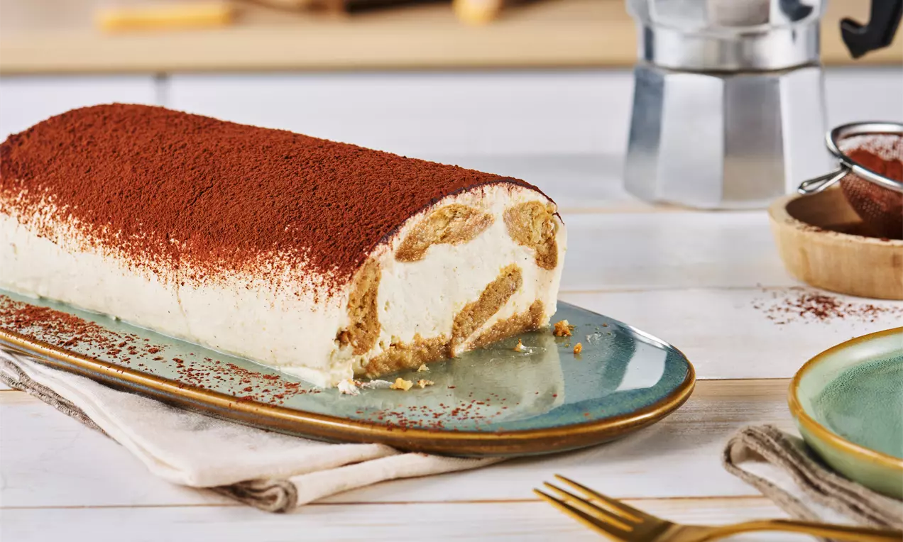 Cremeweiße Tiramisu-Rolle mit goldbraunen Biskuitstücken, fein bestäubt mit Kakao auf einer türkisfarbenen Platte mit Stoffunterlage