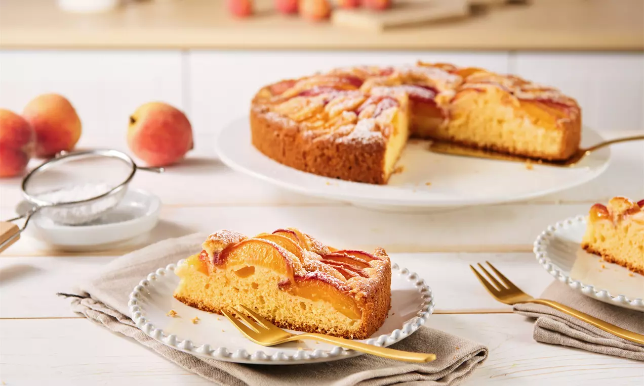 Pfirsichkuchen mit goldgelbem Rührteig und saftigen Pfirsichspalten, leicht mit Puderzucker bestäubt, auf Tellern und einer Tortenplatte angerichtet