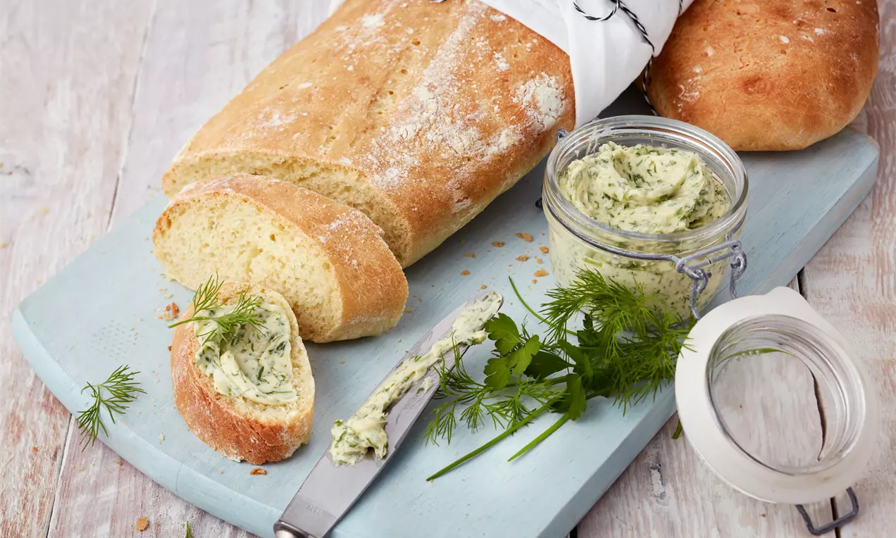 Goldbraunes Ciabatta mit mehliger Kruste, angerichtet mit cremiger Kräuterbutter in einem Glas und frischem Dill auf einem hellblauen Brett