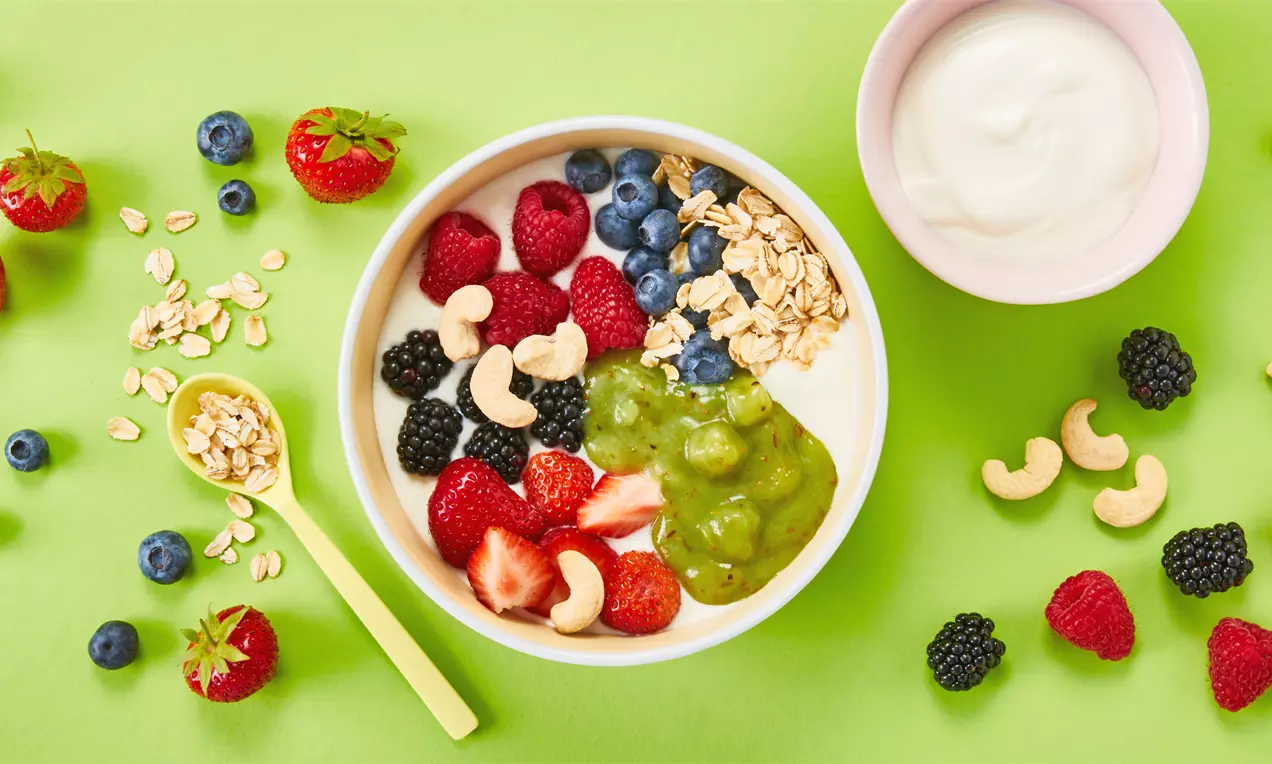 In einer weißen Schale Joghurt mit roten Beeren, Blaubeeren, Cashews, Granola und grünem Fruchtpüree, dekoriert auf grünem Untergrund