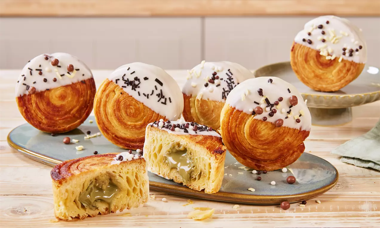 Ttrendige New York Rolls mit goldbrauner Kruste, teils weiß glasiert und bestreut, auf Keramikteller angerichtet, eines mit Pistaziencreme im Kern geöffnet