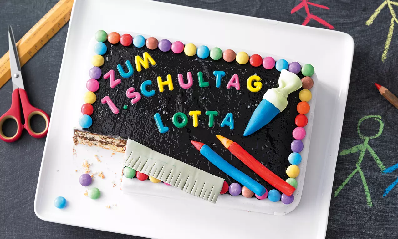 Schokoladen-Schultafel mit buntem Schriftzug, verziert mit essbaren Farbkreiden, Lineal und Zuckerkugeln auf einem weißen Teller