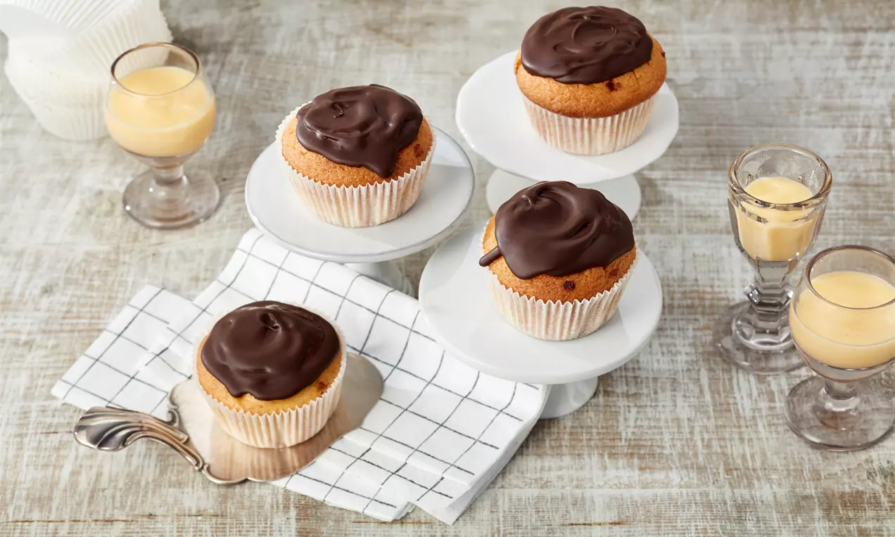 Eierlikör-Muffins mit goldgelbem Teig und glänzender Schokoglasur in weißen Papierförmchen auf gestuften Tellern, daneben Eierlikör in Gläsern