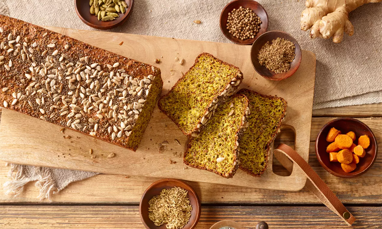 Würziges Eiweißbrot mit gold-grünlicher Krume und grobem Körnermantel, in Scheiben geschnitten auf einem Holzbrett mit Gewürzschälchen drapiert