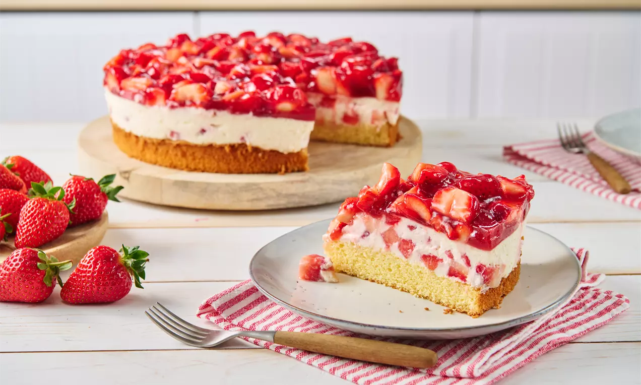 Erdbeerkuchen mit roter Fruchtschicht auf weißer Quarkcreme, gelbem Boden und frischen Erdbeeren auf einem Teller mit Gabel