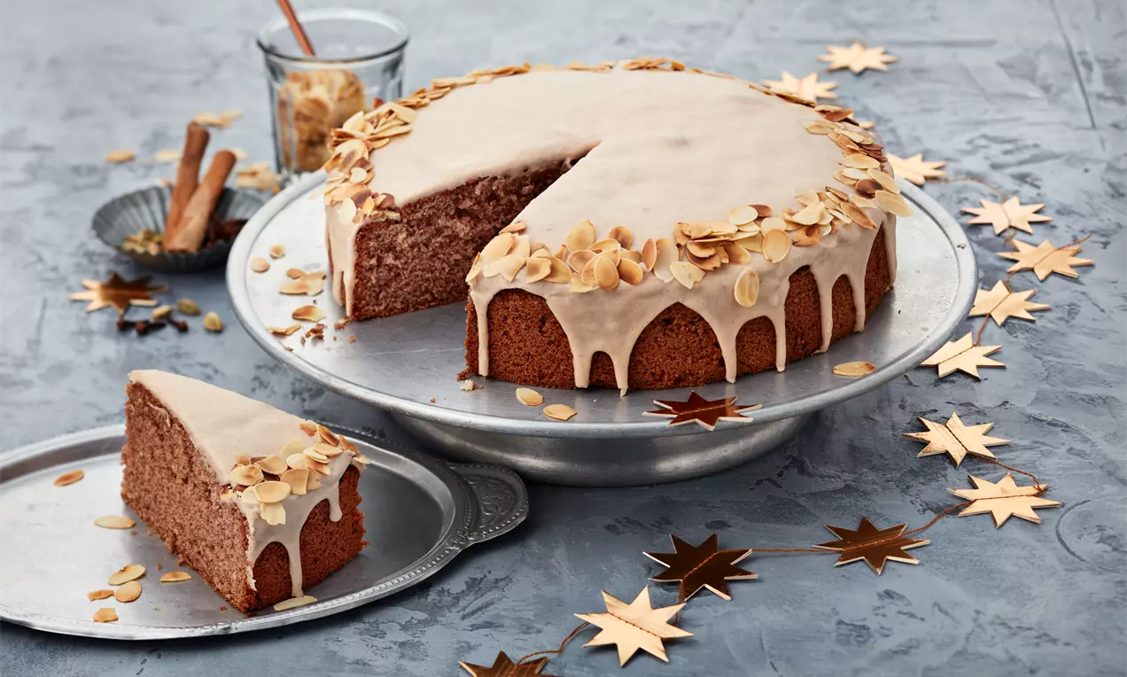 Runder brauner Gewürzkuchen auf silberner Platte, mit heller Glasur und goldbraunen Mandelblättchen, umgeben von Stern-Dekoration