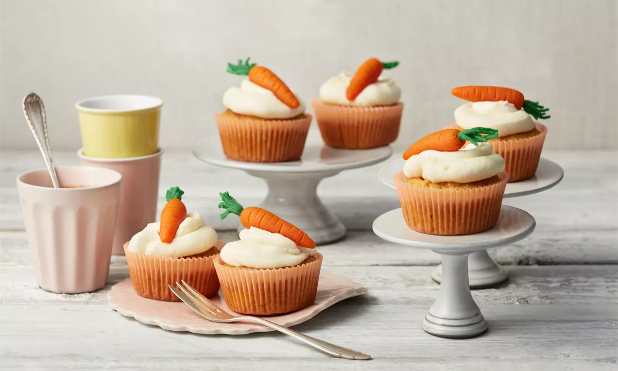 Rübli-Frischkäse-Cupcakes mit orangefarbenen Förmchen und weißer Creme, garniert mit Marzipanmöhren, präsentiert auf weißen Kuchenplatten