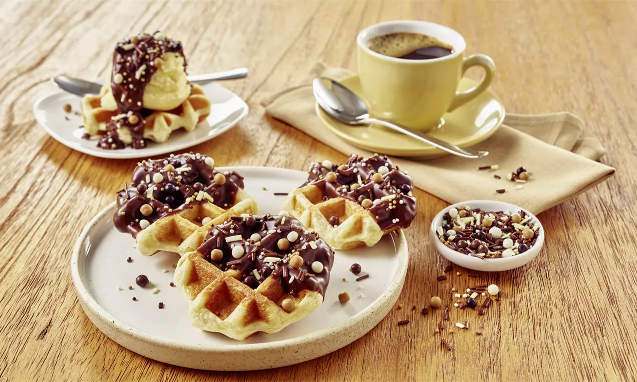 Goldbraune Belgische Waffeln mit Schokoladenglasur und Streuseln auf einem weißen Teller, neben einem gelben Kaffeetasse auf Holzplatte