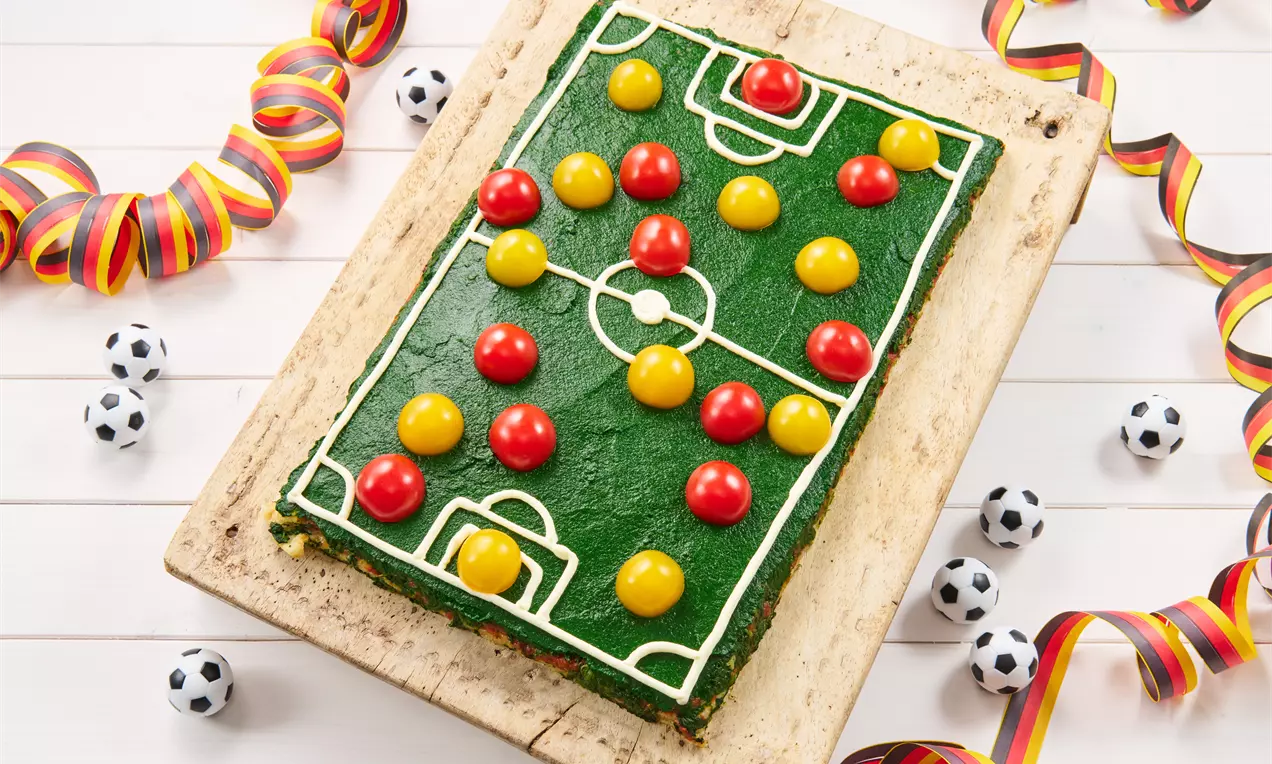 Grüne rechteckige Pizza mit Fußballfeld aus weißer Glasur, gelben und roten Belägen, dekorativ umrahmt von Bändern und Miniaturbällen