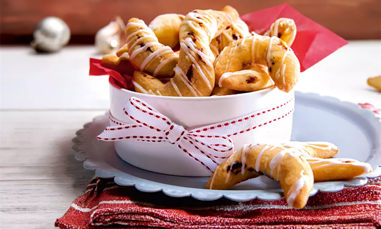 Gebogene Kipferl mit goldbrauner Kruste und dunklen Rosinen, dekorativ mit Zuckerguss verziert in einer weißen Schale mit rotem Band
