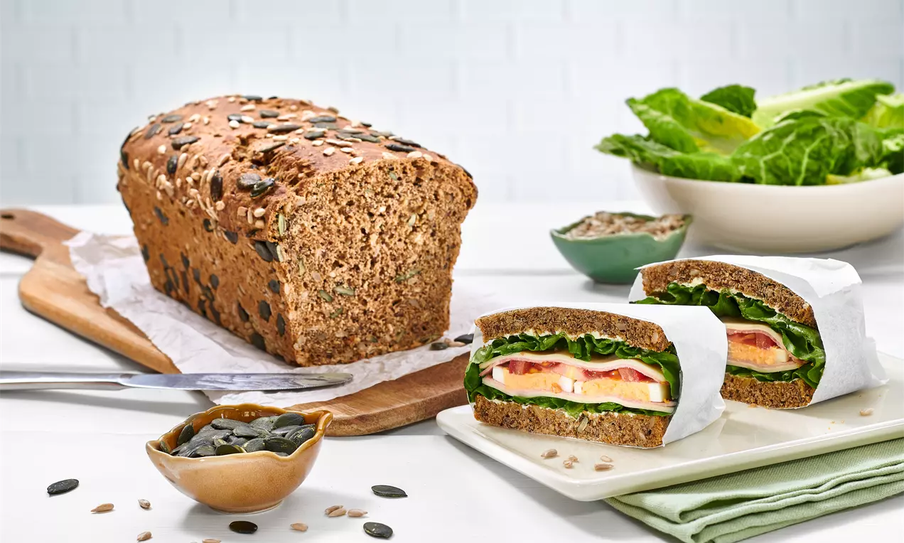 Körniges Vollkornbrot mit dunkler Kruste und Kernen, aufgeschnitten mit Sandwichhälften, gefüllt mit Salat, Käse und Tomaten, neben Kürbiskernen und Salatblättern auf hellem Untergrund drapiert