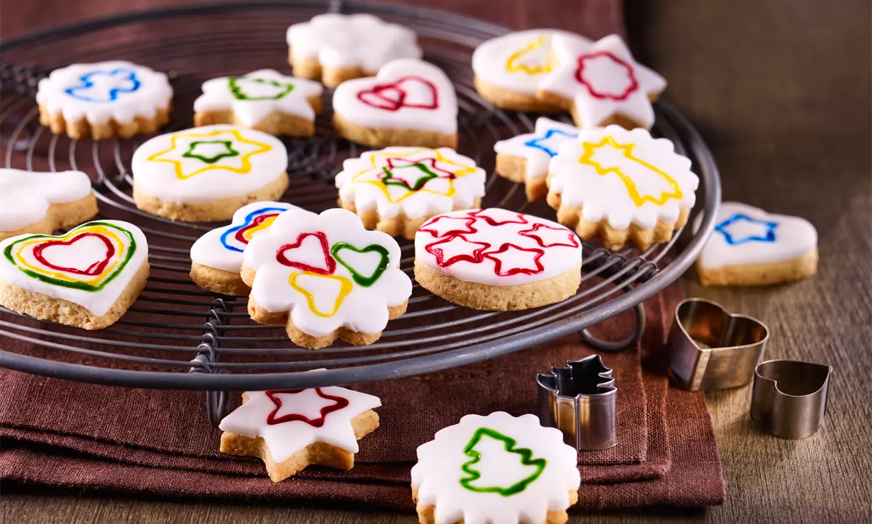 Fondant-Plätzchen in verschiedenen Formen mit bunter Zuckerdekoration auf einem runden Kuchengitter, arrangiert auf brauner Stoffunterlage