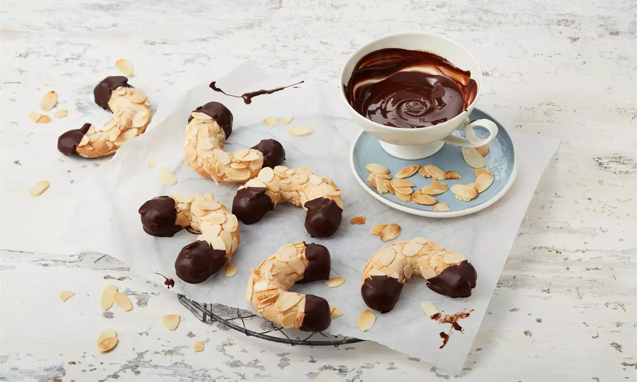 Kleine gebogene Mandelhörnchen mit goldenen Mandelblättchen und dunklen Schokoladenspitzen auf Backpapier neben einer Tasse Schokolade