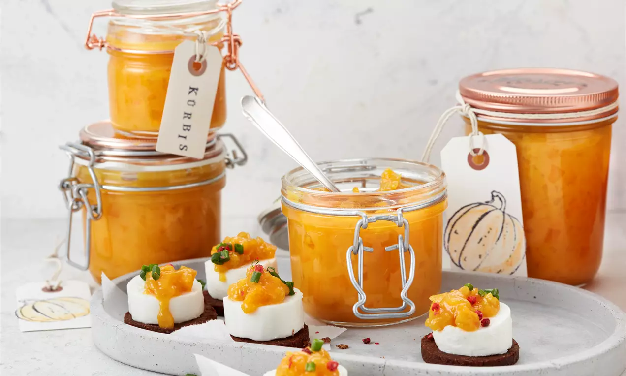 Helles, orangefarbenes Kürbis-Chutney in Gläsern mit Metallverschluss, serviert auf einer grauen Platte mit Cracker und Frischkäse