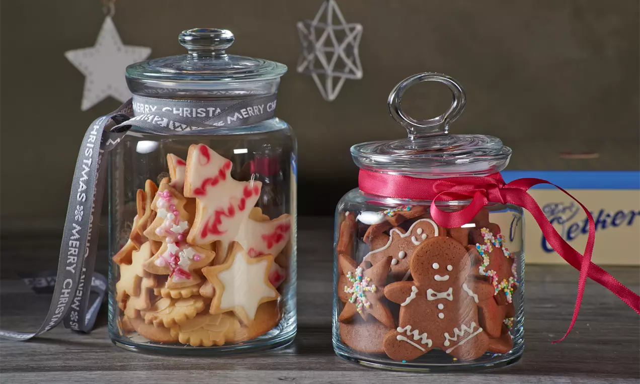 Zwei Glasgefäße mit Deckeln, gefüllt mit bunt verzierten Weihnachtsplätzchen, dekoriert mit Schleifen auf einem Holztisch vor dunklem Hintergrund
