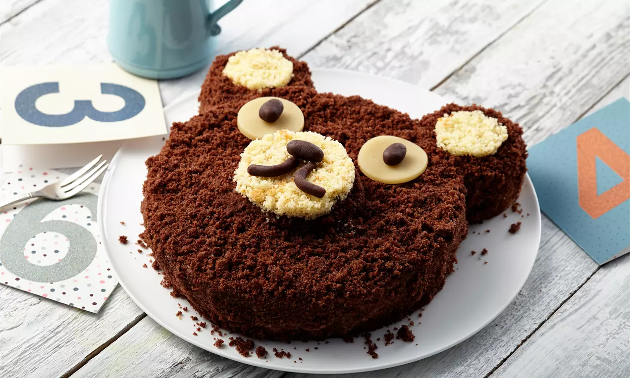 Schokoladiger Bärenkuchen in Form eines Teddykopfs mit heller Creme, Schokodekoration und braunen Krümeln auf weißem Teller angerichtet