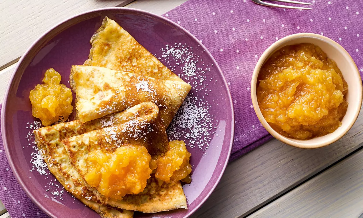 Zarte Pfannkuchen im Dreieck gefaltet, mit Quitten-Orangen-Konfitüre und Puderzucker auf lilafarbenem Teller mit passendem Stoffläufer