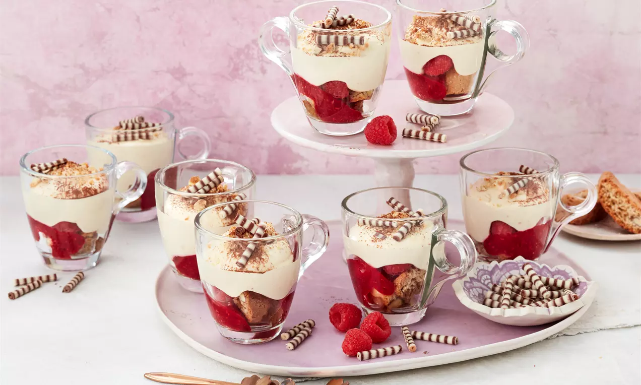 Dessert aus heller Creme auf geschichtetem roten Fruchtpüree, garniert mit Schokospänen in Glastassen auf rosa Platte mit Himbeeren