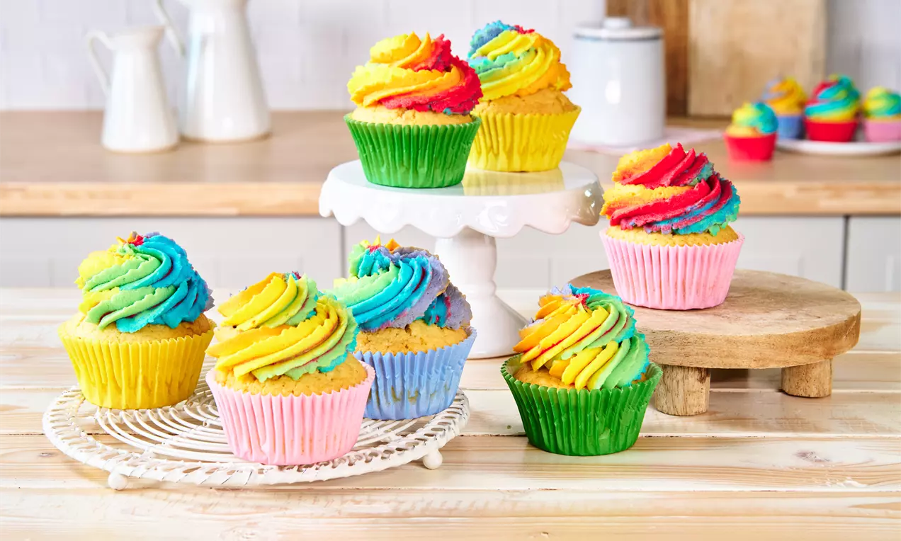 Regenbogenfarbene Cupcakes mit geschwungener, bunter Cremehaube auf grünen und rosa Förmchen, arrangiert auf Holz- und weißen Unterlagen