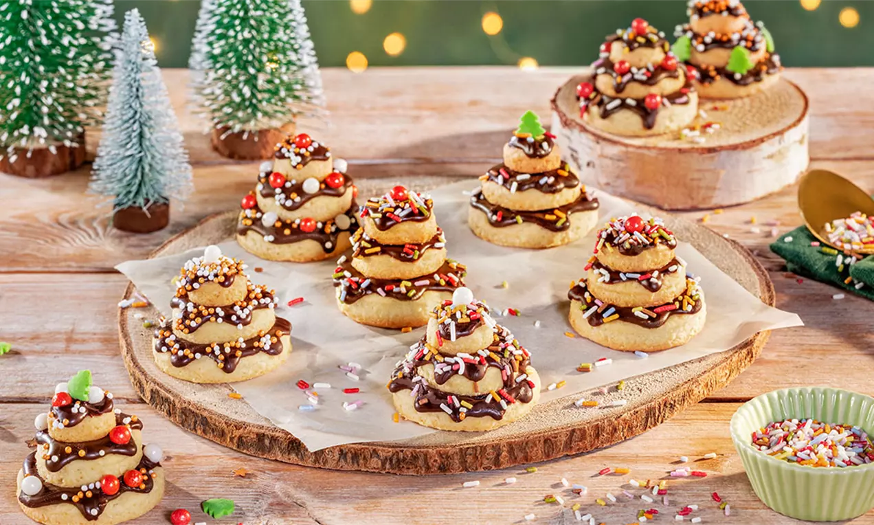 Drip-Cake-Plätzchen in Form kleiner Tannenbäume aus gestapelten Keksen mit dunkler Glasur, bunten Streuseln und Deko auf Holzplatte und Papier